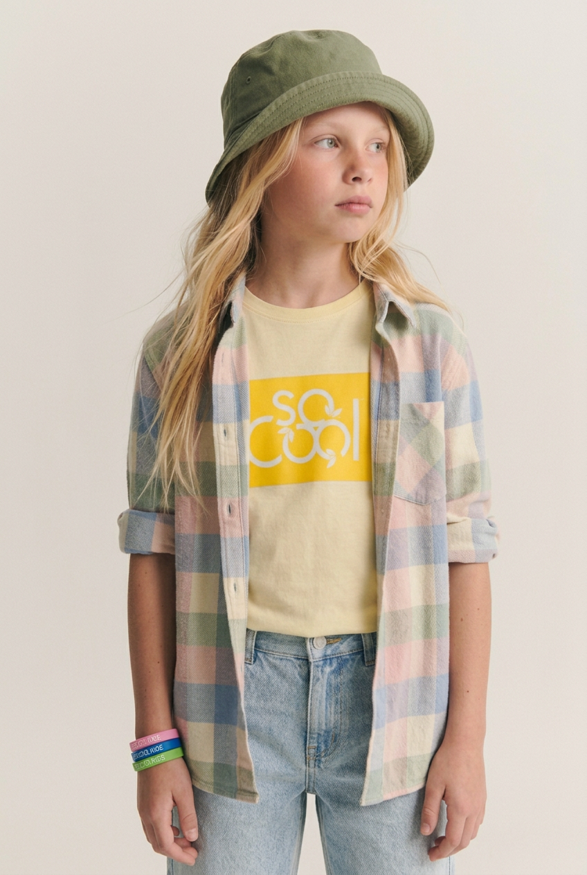 Girl in an olive bucket hat wearing the yellow logo tee, pastel flannel and bracelets